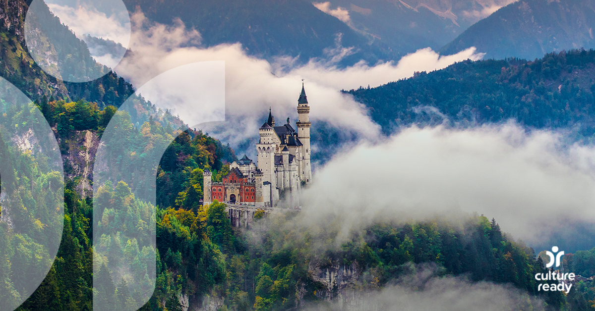 A Large brick and stone castle is nestled between mountains covered in dark green trees with with clouds in the foreground