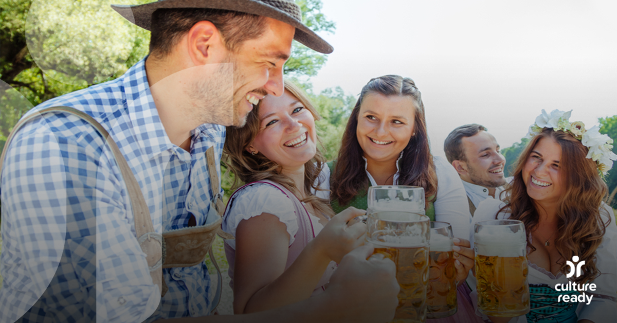 Five people in traditional Bavarian outfits are drinking steins of beer outdoors.