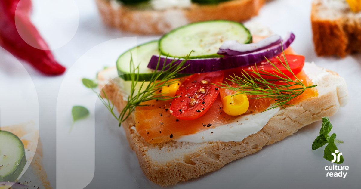 A slide of white bread topped with a white spread, salmon, tomatoes, cucumber, corn, red onion and dill on a white table