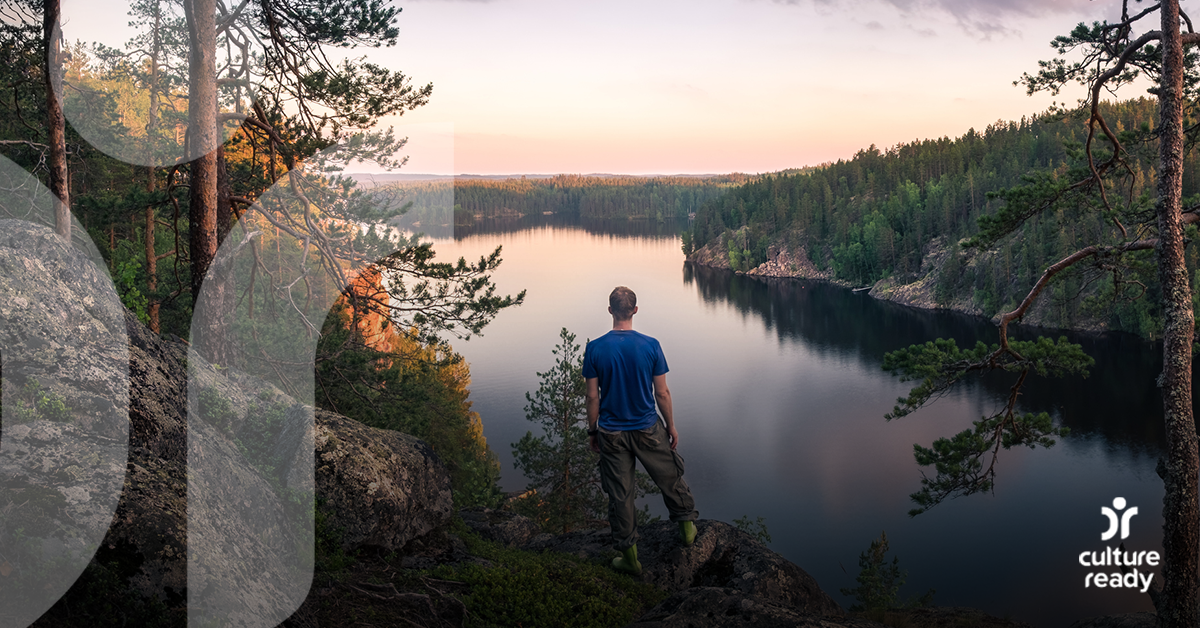 A man in a blue shirt stands on a rock overlooking a river surrounded by evergreen trees during sunrise