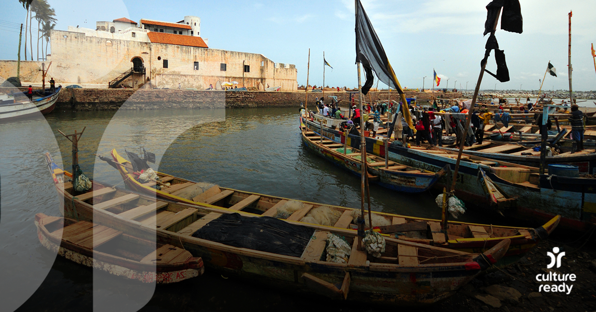 Several wooden boats float on calm water in the foreground, with people gathering on one boat. In this distance is a neutral colored concrete building on a pier. 