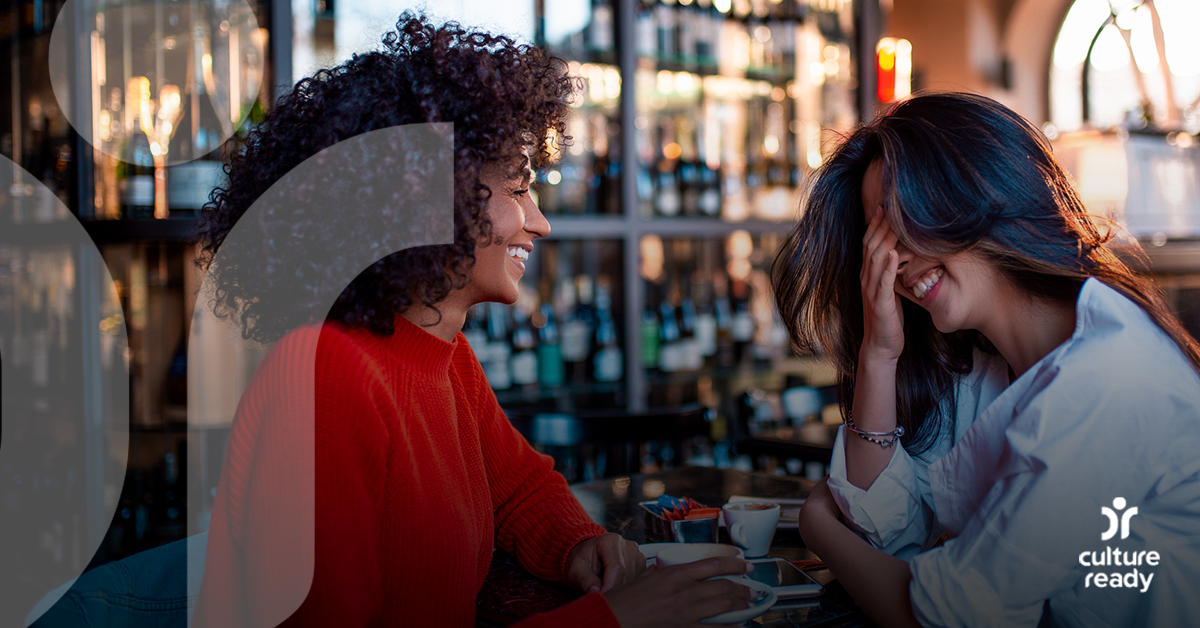 Two women, one with dark curly hair wearing red and one with straight hair wearing white, are laughing while sitting at an open air bar