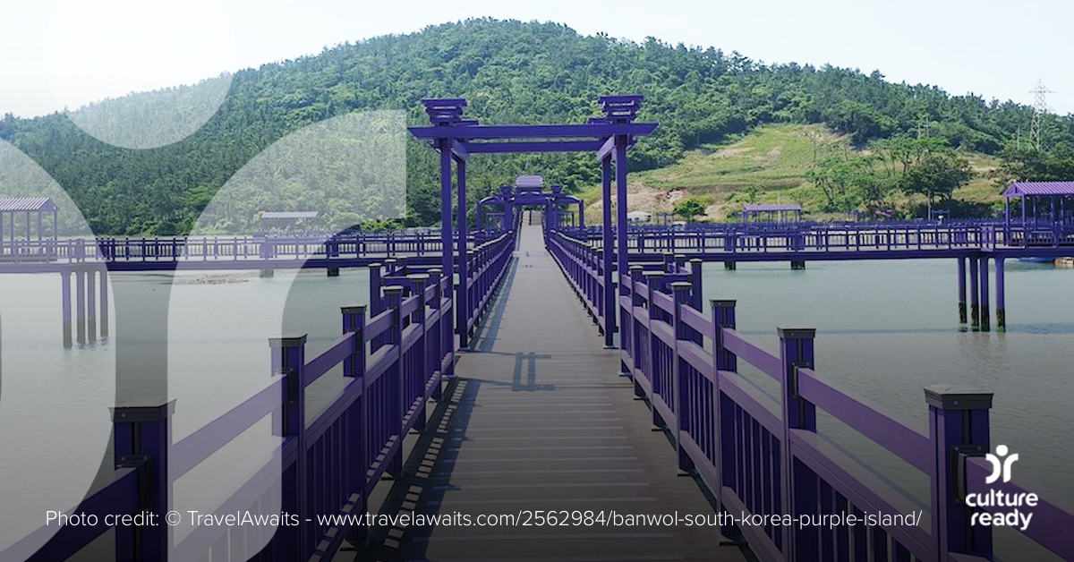 A vibrantly painted purple bridges stretches into the distance over calm water, disappearing into a green hill