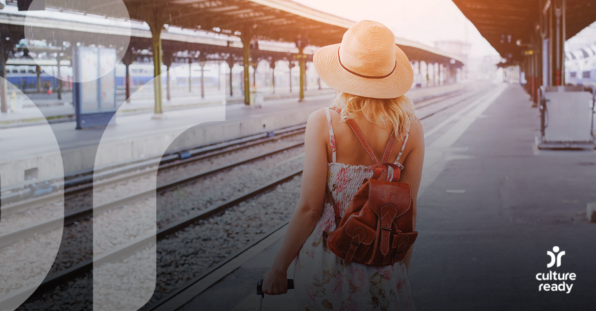 A woman in a straw hat and carrying a brown leather backpack stands on a empty train platform looking into the horizon