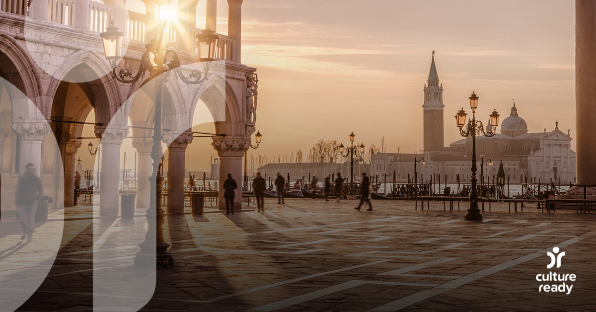 An Italian city square at sunset with stone buildings in the foreground and a large church and clock tower in the background