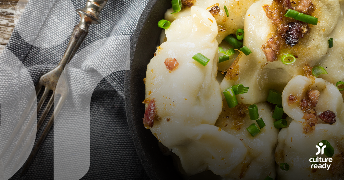 A close up image of a black bowl full of steamed pierogi topped with scallions sits on a gray napkin and silver fork