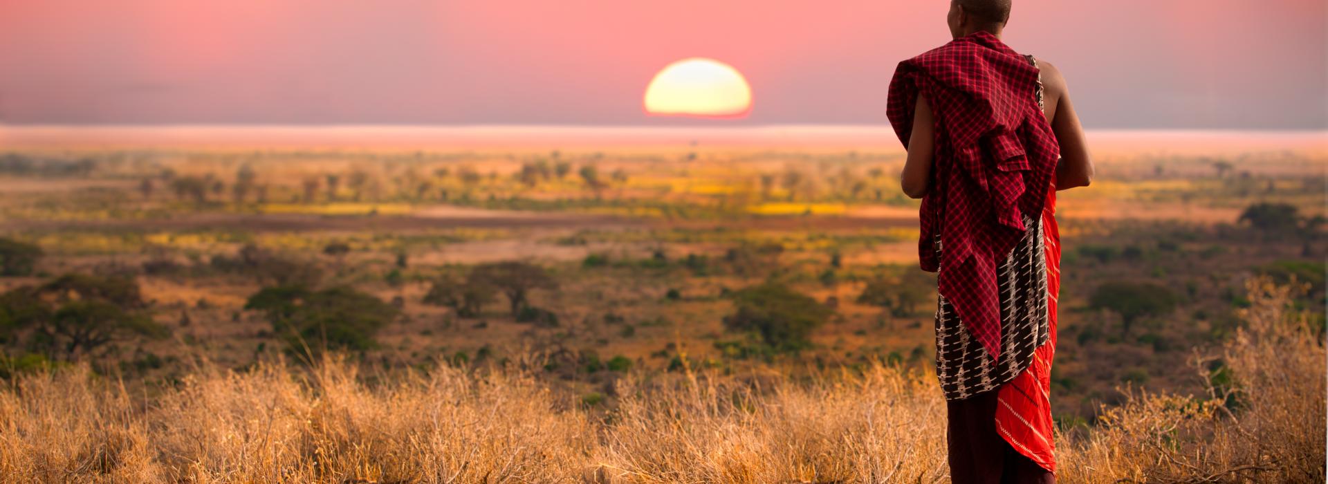 Man wearing colorful fabric walking barefoot on grass towards sunset.